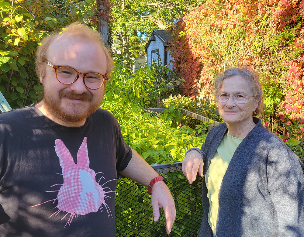 a photo of a man with a red beard and glasses standing next to a woman with grey hair and glasses, in an overgrown garden